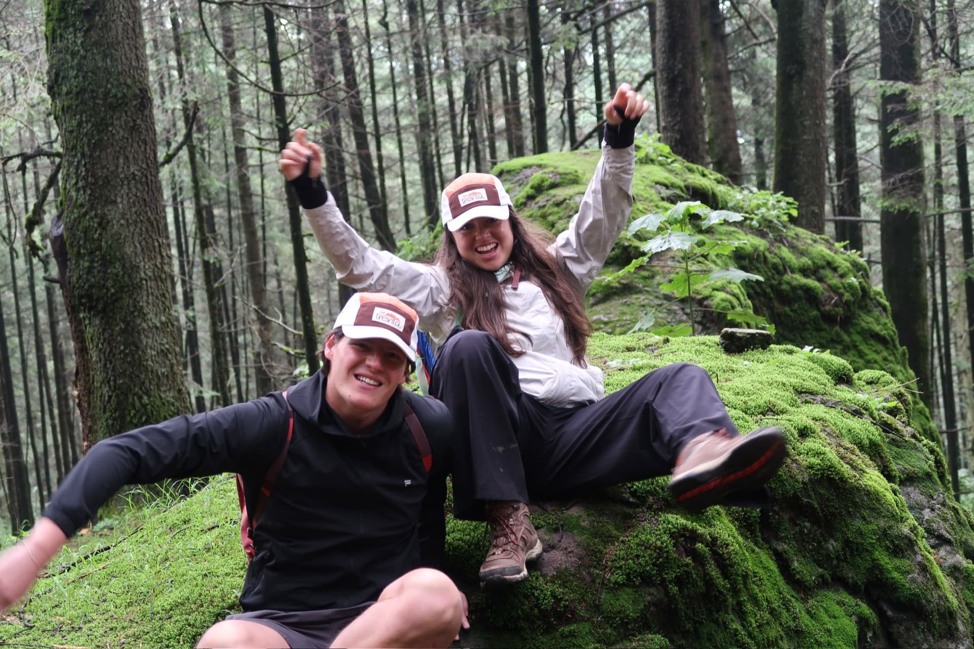 Two people wearing the cap, arms up, on a mossy rock